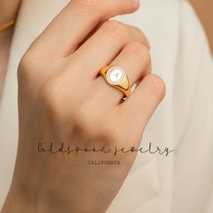 Puede incluir: Un anillo de sello de oro con una piedra ovalada blanca engastada con pequeñas piedras transparentes alrededor del borde. La piedra está grabada con la letra "A".
