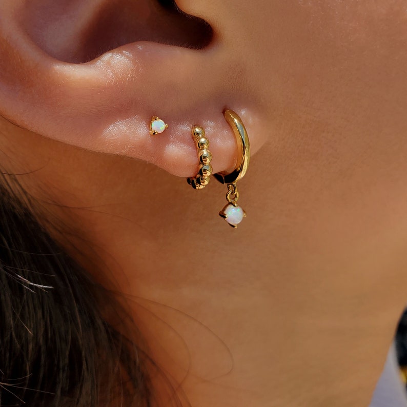 May include: Gold hoop earrings with a small opal dangling from the bottom. The hoop is made of small gold beads. A small opal stud earring is also visible in the image.