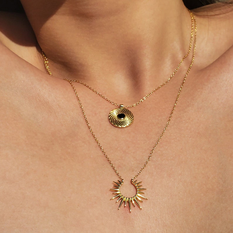 Op de afbeelding: Twee gouden kettingen met hangers. De bovenste ketting heeft een ronde hanger met een zwart midden en gouden draaiende lijnen. De onderste ketting heeft een zonnestralenhanger met een gouden cirkel en puntige stralen.
