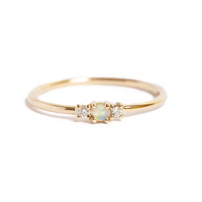 May include: A delicate gold ring featuring a central opal stone flanked by two smaller, clear gemstones. The ring has a thin band and a simple, elegant design, perfect for everyday wear. The gemstones are set in a prong setting.