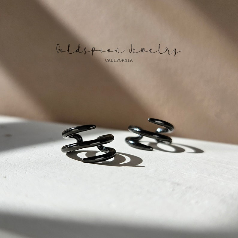May include: Two black metal ear cuffs with a triple spiral design. The ear cuffs are on a white surface. Goldspoon Jewelry California.