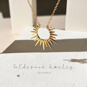 May include: A gold-colored necklace with a sunburst pendant. The pendant features a circular center with radiating spikes. The necklace is displayed on a white surface, with the text "Goldspoon Jewelry CALIFORNIA" at the bottom.