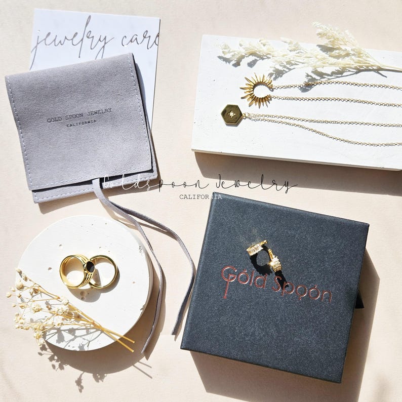 Puede incluir: Una colecci&oacute;n de joyer&iacute;a dorada que incluye dos collares con colgantes de sol y hexagonales, dos anillos con incrustaciones negras y dos peque&ntilde;os pendientes de aro. Los art&iacute;culos se muestran con una bolsa de ante gris y una caja negra.