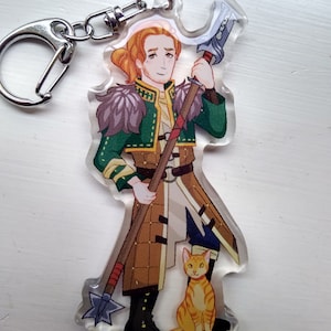 Peut inclure: Un porte-clés représentant une illustration de personnage de dessin animé avec de longs cheveux roux tenant une lance avec une pointe en forme d'étoile. Le personnage porte une tenue verte et marron et se tient à côté d'un petit chat orange.