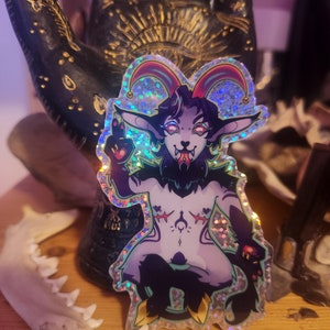 May include: A holographic sticker of a purple and black creature with horns, wearing a jester hat. The creature has a tongue sticking out and is making a peace sign with its hand.