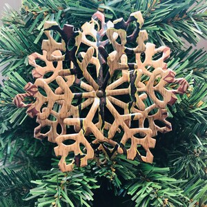 May include: A wooden snowflake ornament with a natural wood grain finish and brown and red accents. The snowflake is attached to a green Christmas tree. The ornament has a rustic, festive appearance.