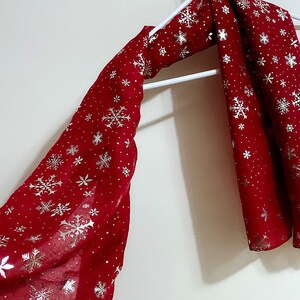Puede incluir: Una bufanda roja con un patr&oacute;n de copo de nieve en plata. La bufanda est&aacute; colgada de una percha blanca.