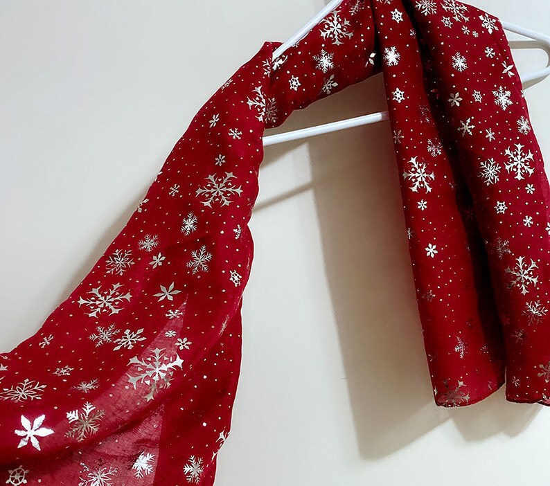 Puede incluir: Una bufanda roja con un patr&oacute;n de copo de nieve en plata. La bufanda est&aacute; colgada de una percha blanca.