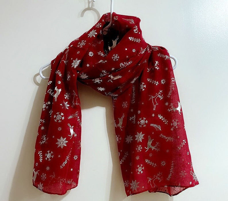 Puede incluir: Una bufanda roja de Navidad con un patr&oacute;n de copo de nieve y reno plateado. La bufanda tiene la palabra "Navidad" escrita varias veces.