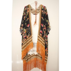 Op de afbeelding: Een zwarte bloemenkimono met oranje franjes. De kimono heeft een geometrisch patroon en is gemaakt van een lichtgewicht stof.