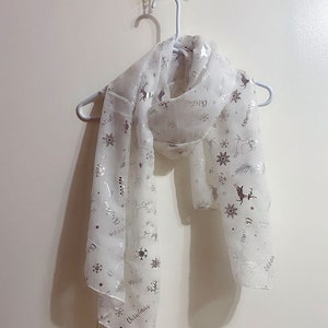 Puede incluir: Bufanda blanca transparente con un patr&oacute;n de copo de nieve y reno plateado. La bufanda tiene la palabra "Christmas" impresa.