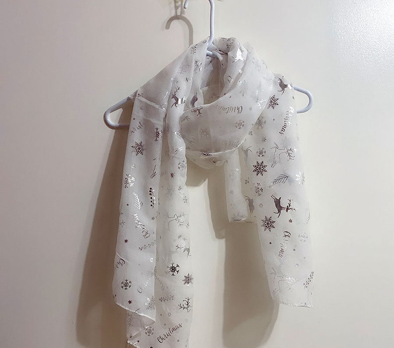 Puede incluir: Bufanda blanca transparente con un patr&oacute;n de copo de nieve y reno plateado. La bufanda tiene la palabra "Christmas" impresa.