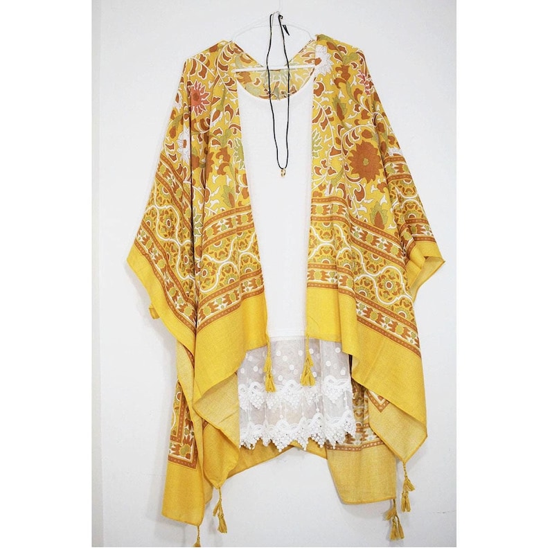 K&ouml;nnte beinhalten: Ein gelbes und braunes gemustertes Kimono mit wei&szlig;em Spitzenbesatz und Quasten. Das Kimono ist vorne offen und hat ein wei&szlig;es Tanktop darunter.