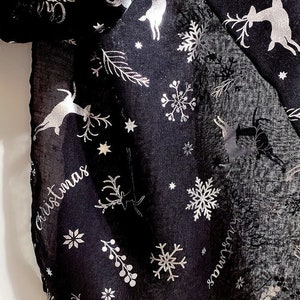 Puede incluir: Bufanda negra de gasa con estampado met&aacute;lico plateado de renos, copos de nieve y estrellas. La bufanda tiene la palabra "Christmas" escrita en letras met&aacute;licas plateadas.