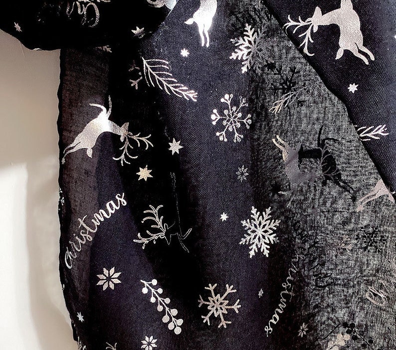 Puede incluir: Bufanda negra de gasa con estampado met&aacute;lico plateado de renos, copos de nieve y estrellas. La bufanda tiene la palabra "Christmas" escrita en letras met&aacute;licas plateadas.