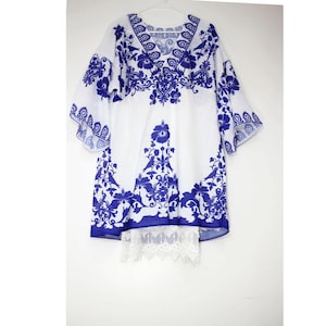 Puede incluir: Vestido túnica blanco con estampado floral azul y ribete de encaje. El vestido tiene un escote en V y mangas 3/4.