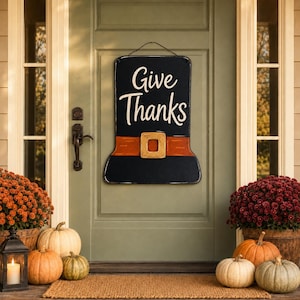 Puede incluir: Un letrero de pared negro con forma de sombrero que presenta el texto blanco "Give Thanks" sobre un cinturón naranja con una hebilla dorada. El letrero está suspendido por un alambre fino y diseñado para colgarse en una puerta con estilo festivo.