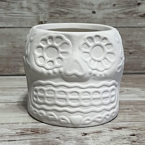 Puede incluir: Una maceta de cerámica blanca con forma de calavera de azúcar, con intrincados patrones florales y geométricos. La maceta tiene una abertura ancha y está diseñada para contener plantas u otros objetos decorativos. El diseño de la calavera es un tema popular para las celebraciones del Día de Muertos.