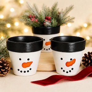 Peut inclure: Trois pots en céramique blanche avec des bords noirs, chacun peint avec un visage de bonhomme de neige comprenant un nez orange en forme de carotte et des yeux et une bouche en points noirs. Les pots sont disposés avec deux devant et un derrière.