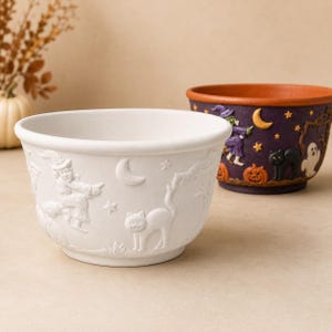 May include: A white ceramic bowl featuring embossed Halloween motifs including a witch on a broomstick, a crescent moon, stars, and a cat. Behind it sits a second bowl in dark purple with colorful painted details of a witch, cat, and ghost.