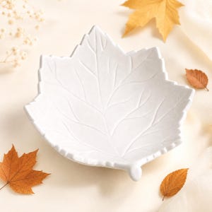 May include: A white, leaf-shaped dish with a matte finish and embossed vein details. The bowl features a pointed tip and a small stem at the base, mimicking the natural structure of a maple leaf with its distinct serrated edges and central spine.