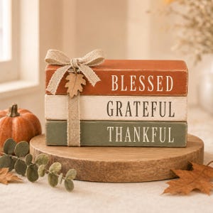 Könnte beinhalten: Ein Stapel aus drei rechteckigen Holzblöcken in gebranntem Orange, Creme und Salbeigrün. Die Blöcke tragen die weißen Schriftzüge BLESSED, GRATEFUL und THANKFUL. Ein beiges Juteband ist um den Stapel gebunden und mit einem kleinen Holzblatt verziert.
