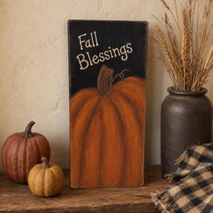 May include: A rectangular wooden sign with a black background and a painted orange pumpkin at the bottom. The text Fall Blessings is written in a cream-coloured script font at the top. The sign has a distressed finish with visible wood grain texture.