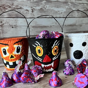 Könnte beinhalten: Drei Halloween-Süßigkeiteneimer: ein orangefarbener Kürbis, eine schwarze Katze und ein weißer Geist. Jeder Eimer hat einen schwarzen Drahtgriff und ist mit lila folienverpackten Süßigkeiten gefüllt. Die Eimer stehen auf einer Holzoberfläche.