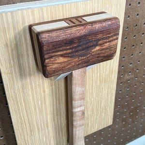 May include: A wooden mallet with a brown, red, and white striped handle. The mallet head is made of dark wood with a light wood inlay.