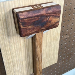 May include: A wooden mallet with a dark brown head and a lighter brown handle. The head features a decorative stripe and the word "Founder" is visible. The mallet is mounted on a light-colored wooden board.