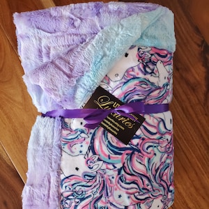 May include: A soft, purple and blue minky baby blanket with a white cotton fabric featuring a unicorn print. The blanket is tied with a purple ribbon and has a tag that reads "Little Minky Luxuries".