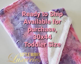 May include: A soft, plush toddler blanket in shades of pink, purple, and white. The blanket measures 30 x 44 inches and features a textured, faux fur design. The blanket is folded, revealing a white, fluffy interior.