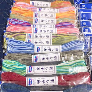 Puede incluir: Una colección de madejas de hilo Sashiko de colores en envases de plástico transparente. Los hilos son de varios colores, incluyendo naranja, rosa, azul, verde y rojo. Cada madeja tiene una etiqueta blanca con texto japonés y la marca "Olympus".