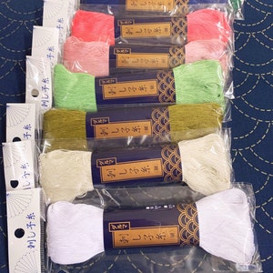Op de afbeelding: Een verzameling kleurrijke borduurgaren spoelen in diverse tinten, waaronder groen, roze en wit. Elke spoel is verpakt in transparant plastic en heeft een label met Japanse tekst. De spoelen zijn netjes gerangschikt.