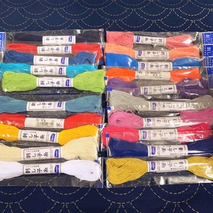 Op de afbeelding: Een verzameling van 20 spoelen borduurgaren in verschillende kleuren, waaronder rood, blauw, groen, geel, oranje, paars, roze en wit. De spoelen zijn verpakt in doorzichtige plastic zakjes met etiketten in het Japans.