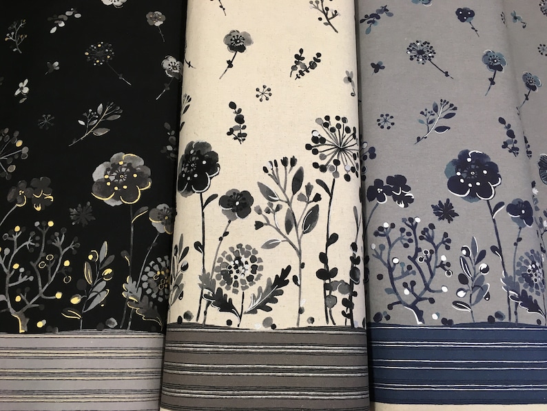 May include: Three rolls of fabric with floral designs. The fabric on the left is black with gold highlights, the middle is off-white, and the right is grey. Each has a border of horizontal stripes.
