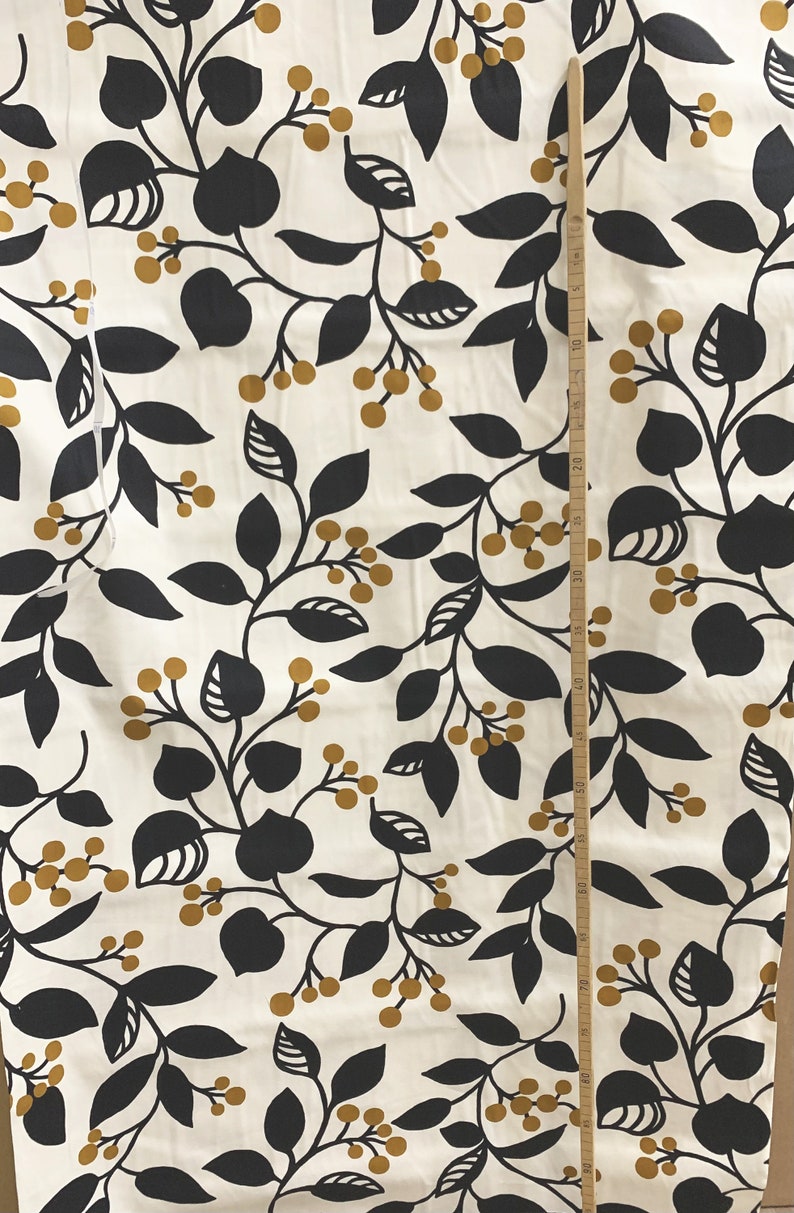 May include: Cream-colored fabric with a repeating pattern of black leaves and golden-brown berries. A wooden ruler is placed vertically along the fabric, showing measurements in inches. The design is suitable for home decor or apparel.