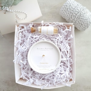 May include: A white gift box filled with white crinkle paper containing a round white soy candle labeled Aroma Harvest Candle Co. Coastal and a small glass vial with a cork stopper filled with wooden matches, alongside a spool of grey and white twine.