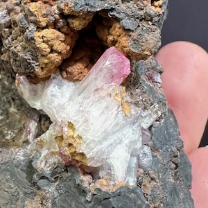 May include: A dark grey rock sample with a rough texture, showing areas of brown and orange. Embedded within the rock are clusters of white and pink crystal formations. The crystals have a translucent appearance.
