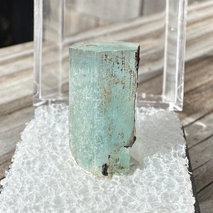 以下が含まれることがあります： 透明なプラスチック製のディスプレイボックスに収められた、薄い青色のアクアマリンの結晶。黒いフォームのベース付き