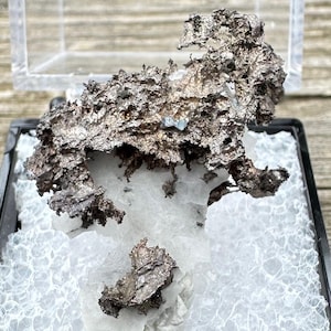 Può includere: Un campione di pepita d'argento in una teca di plastica trasparente con una base di schiuma bianca. La pepita è un ammasso di pezzi metallici irregolari.
