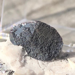 May include: A small, dark grey, metallic mineral specimen with a white, crystalline matrix. The mineral is sitting in a black display box with a white foam lining.