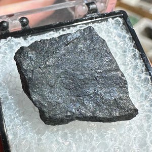 May include: A dark grey mineral specimen, roughly rectangular, presented in a clear plastic case with a black frame. The mineral rests on a bed of white, textured material. The case is open, showing the specimen.