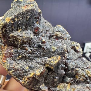 Puede incluir: Un espécimen mineral oscuro y de textura rugosa con una mezcla de tonos negros, marrones y amarillo dorado. Pequeños cristales de color marrón rojizo son visibles en toda la roca. El mineral se sostiene en una mano, mostrando su apariencia natural y sin pulir.