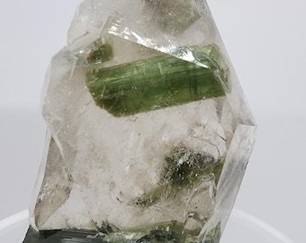 May include: A clear quartz crystal with a green tourmaline inclusion. The crystal is faceted and has a sharp point. The tourmaline is partially visible through the quartz.