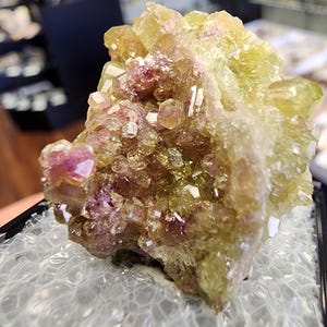 Op de afbeelding: Een close-up van een mineraalmonster met een cluster van kristallen. De kristallen vertonen een mix van kleuren, waaronder lichtgeel, roze en wit, met een ruw, getextureerd oppervlak. Het mineraal wordt gepresenteerd op een helder, webachtig oppervlak.