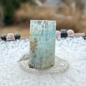 以下が含まれることがあります： 長方形の薄い青色のアクアマリンの結晶が、黒い発泡スチロールで裏打ちされた箱に展示されています