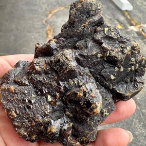 May include: A dark, rough-textured mineral specimen with a mix of black, brown, and gold colors. The rock has a crystalline structure with sharp edges and a natural, unpolished appearance. The mineral is held in a hand.