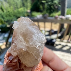 K&ouml;nnte beinhalten: Nahaufnahme eines klaren, durchsichtigen Kristalls mit einem geschichteten, rosa und orangen Sockel. Der Kristall hat eine raue, strukturierte Oberfl&auml;che und wird in einer Hand gehalten.