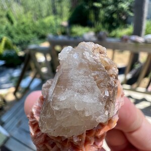 K&ouml;nnte beinhalten: Ein klarer, durchsichtiger Kristallcluster mit einem geschichteten, rosa und orangen Sockel. Der Kristall ist eine nat&uuml;rliche Formation mit einer rauen, strukturierten Oberfl&auml;che.